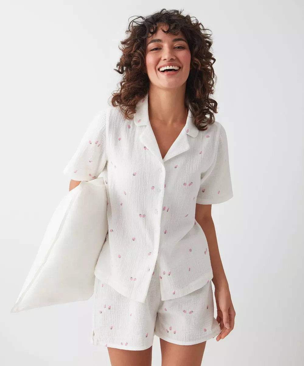 Kobieta w białej piżamie z różowym kwiatowym wzorem, uśmiecha się i trzyma poduszkę.