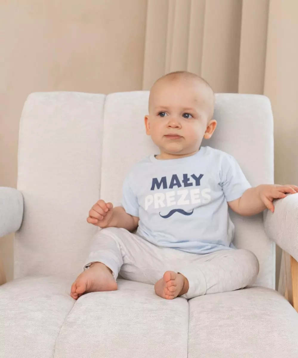 Niemowlę siedzące na beżowym fotelu w niebieskiej koszulce z napisem MAŁY PREZES i beżowych spodniach.