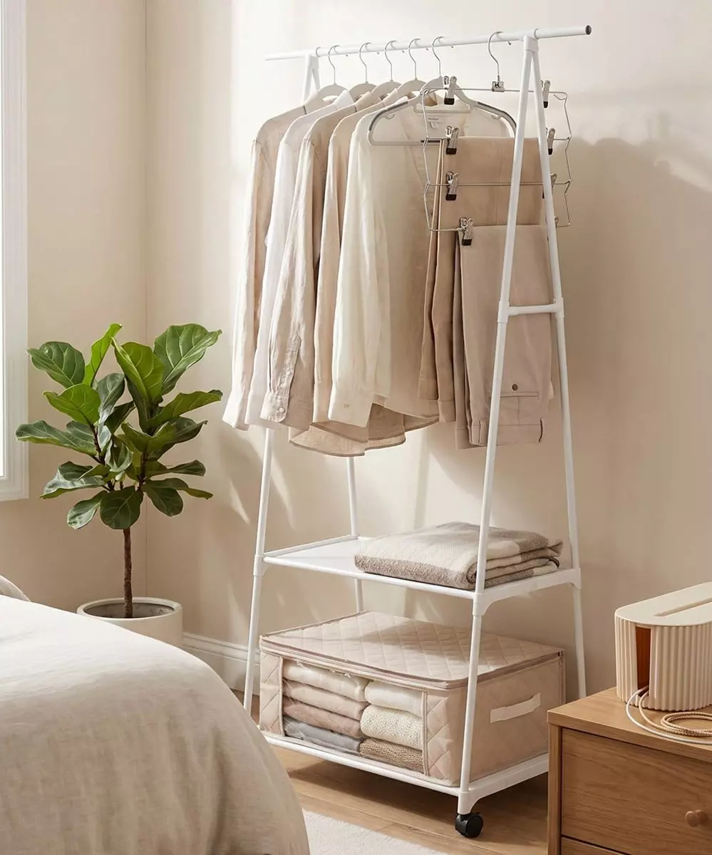 Perchero blanco con ruedas que sostiene ropa en tonos neutros, con estantes inferiores de almacenamiento.