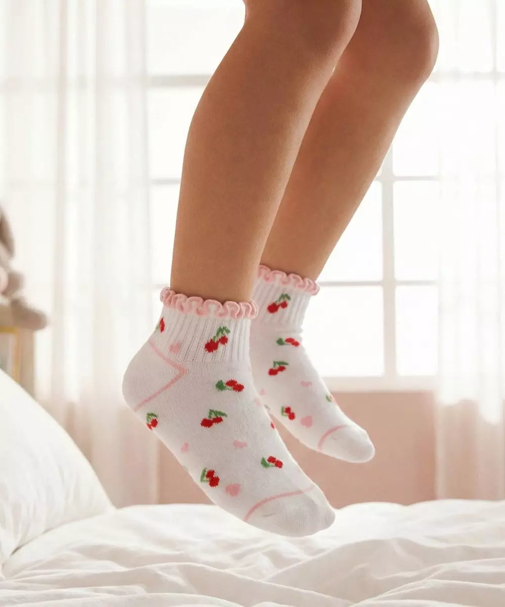 Białe skarpetki z różową falbanką i wzorem w czerwone wiśnie, noszone przez osobę stojącą na łóżku.