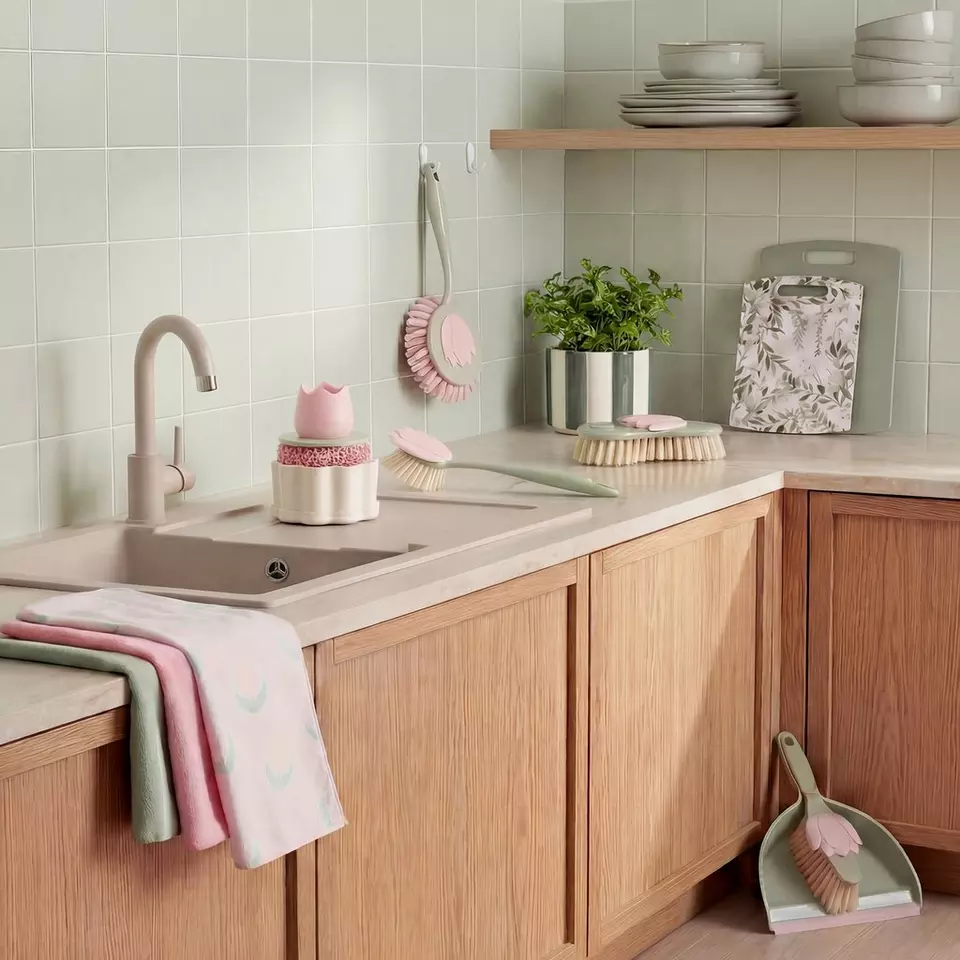 Stylowa kuchnia z drewnianymi szafkami, pastelowymi akcesoriami do czyszczenia i zieloną rośliną w doniczce na blacie.