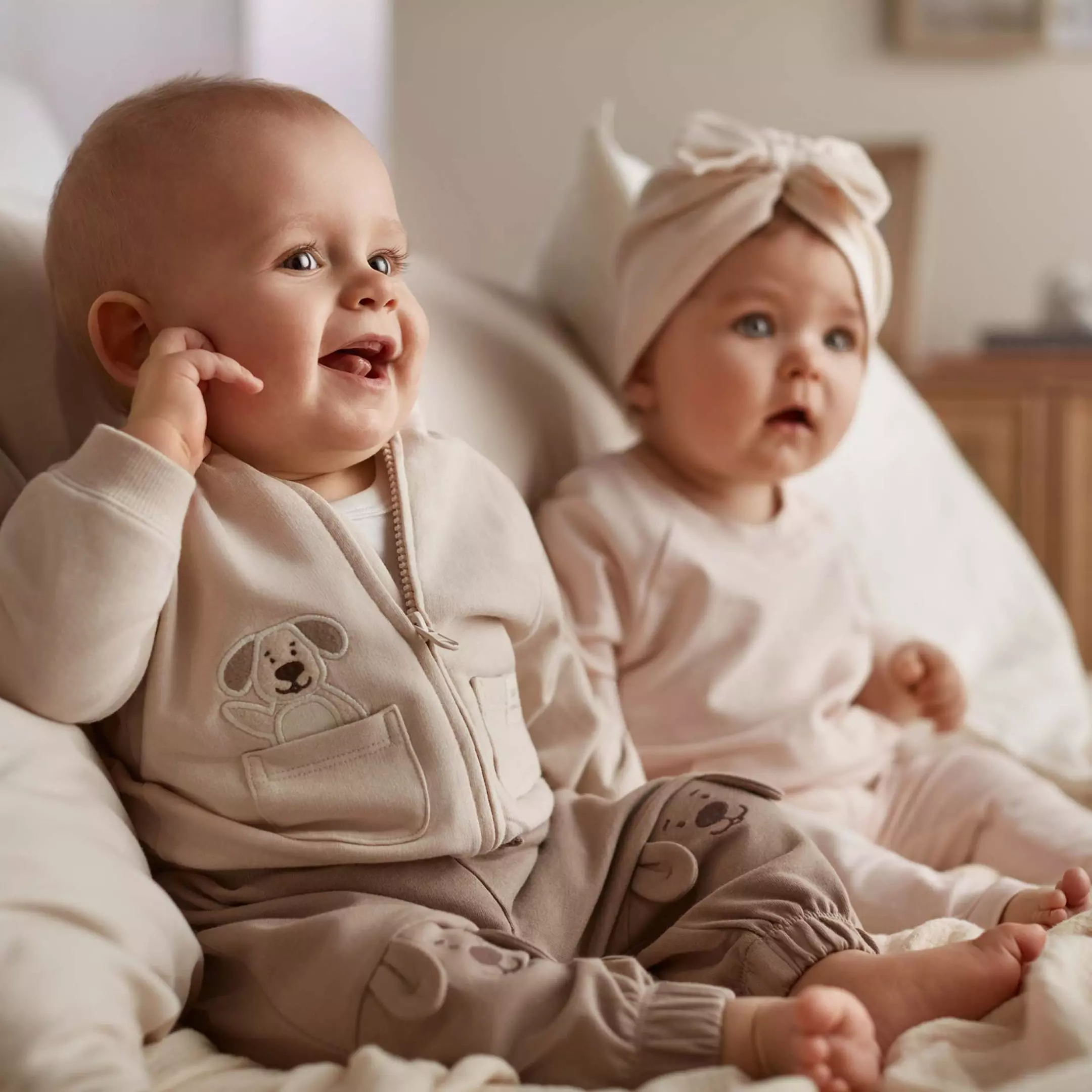 Two babies sitting together wearing beige outfits with puppy designs, one smiling and one wearing a bow headband.