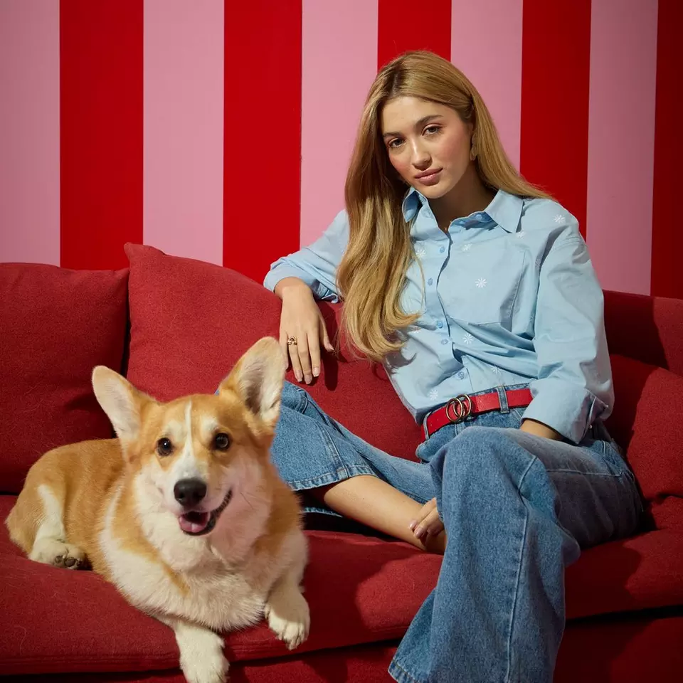Modelka w błękitnej koszuli Pepco z czerwonym paskiem, siedząca na czerwonej kanapie z psem corgi na tle pasiastej ściany.
