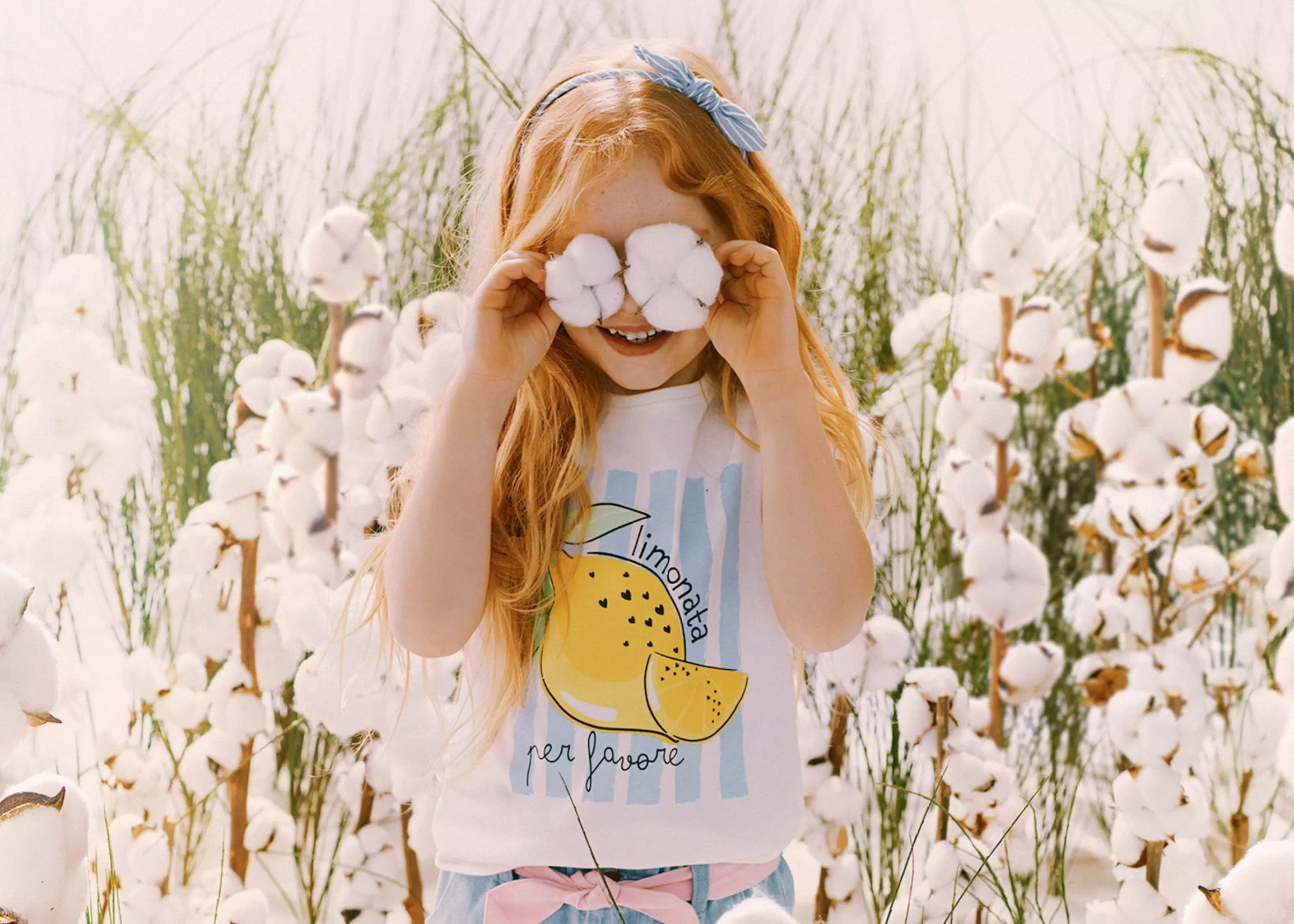 Niña pelirroja con lazo azul sujetando flores de algodón sobre sus ojos en un campo de algodón.