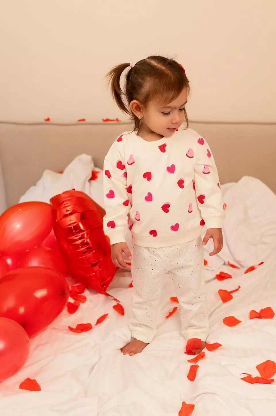 Child wearing Pepco heart-patterned cream sweatshirt and white trousers, standing on bed with red heart balloons and rose petals.