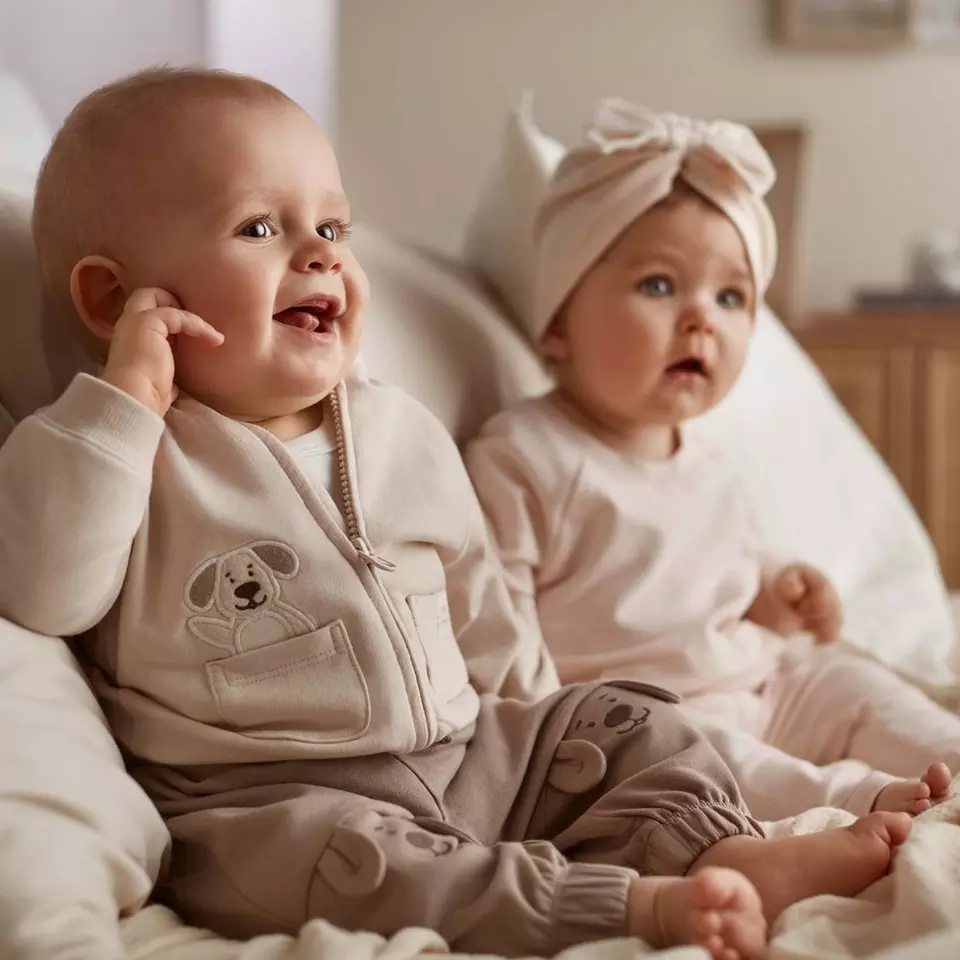 Dwoje niemowląt w beżowych ubraneczkach z aplikacją pieska, jedno dotyka ucha.
