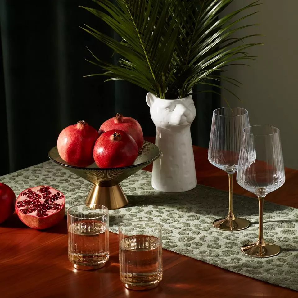 Pepco home decor featuring cat-shaped vase with palm leaves, gold fruit bowl with pomegranates, and elegant wine glasses.