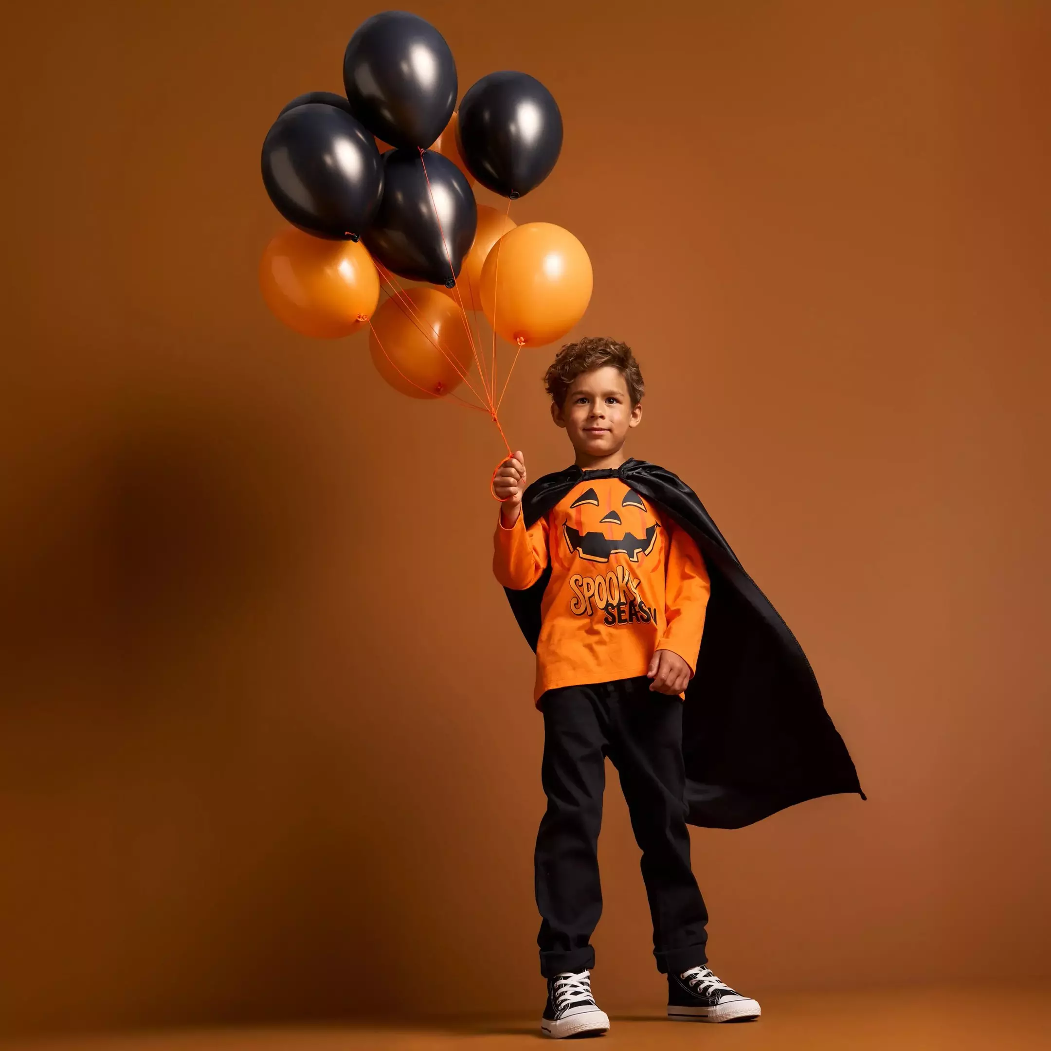 Chłopiec w stroju Halloween Pepco z czarną peleryną trzyma pomarańczowe i czarne balony.