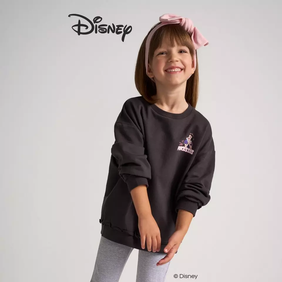 Dziewczynka w czarnej bluzie Pepco z motywem Disney, uśmiechnięta z różową kokardą we włosach.