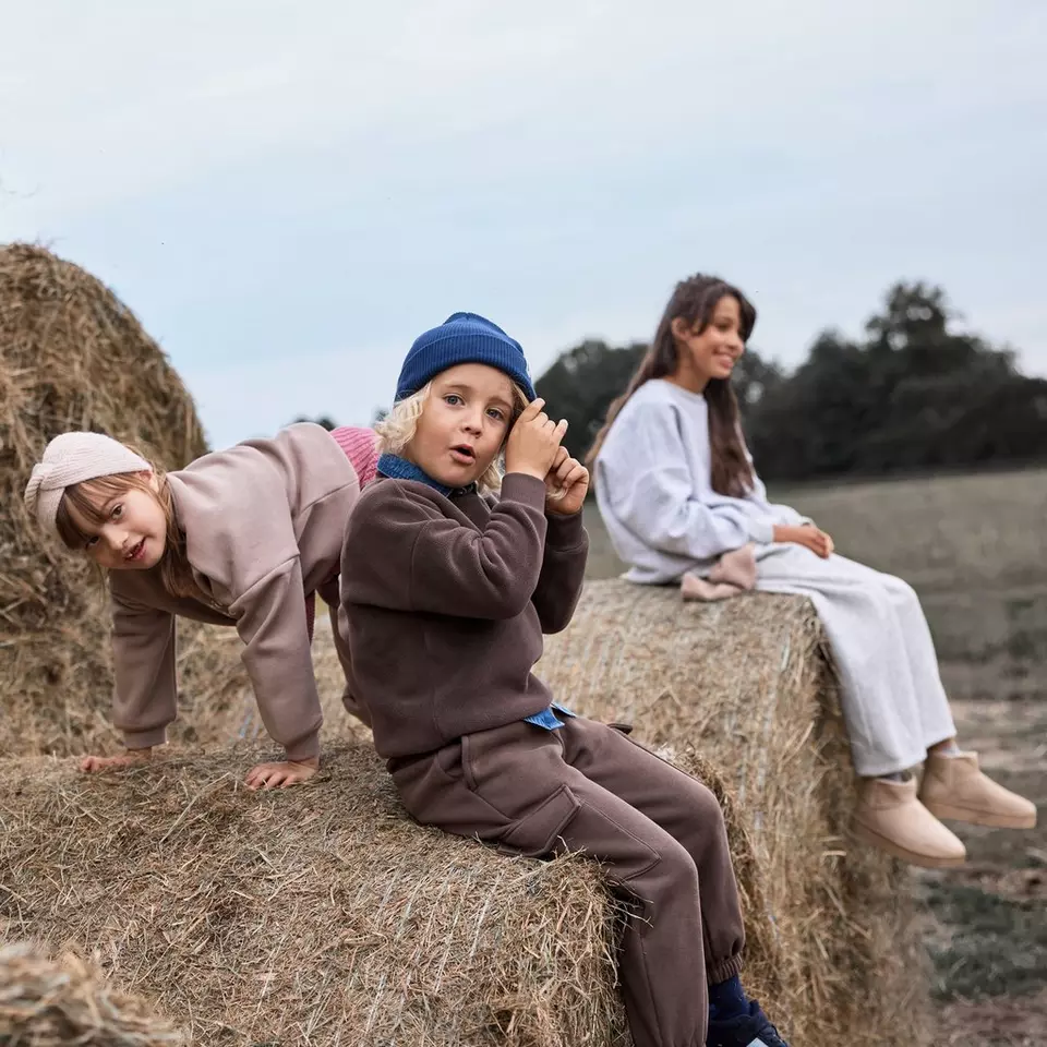 Niños con ropa deportiva Pepco jugando sobre fardos de heno en el campo con una mujer al fondo.