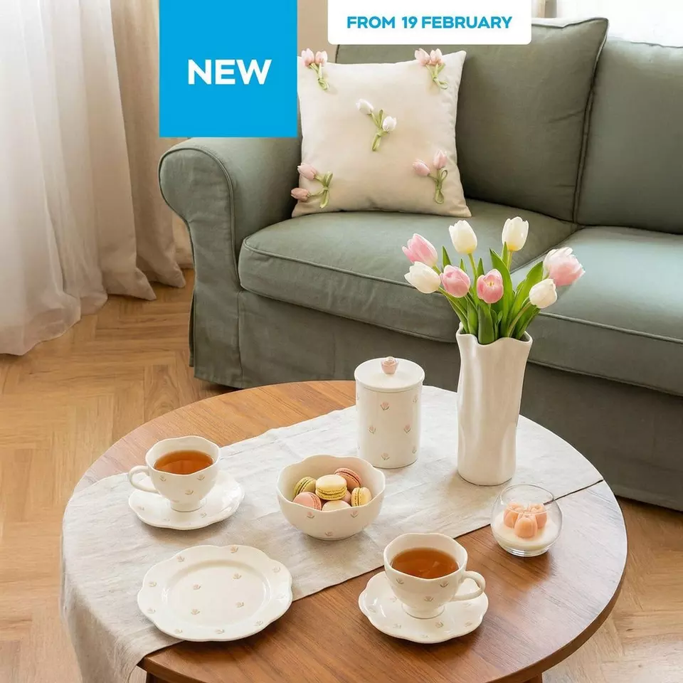 Elegant living room setup with tea cups, macarons and tulips on wooden table. Green sofa with floral cushion. "NEW FROM 19 FEBRUARY" banner.