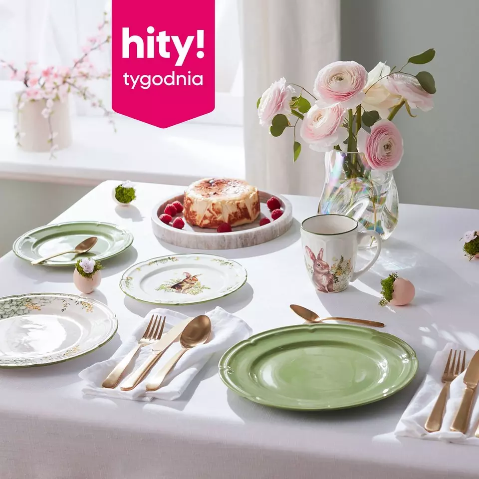 Świąteczny stół z zastawą wielkanocną, sernikiem z malinami, kubkiem z motywem królika i wazonem różowych kwiatów.