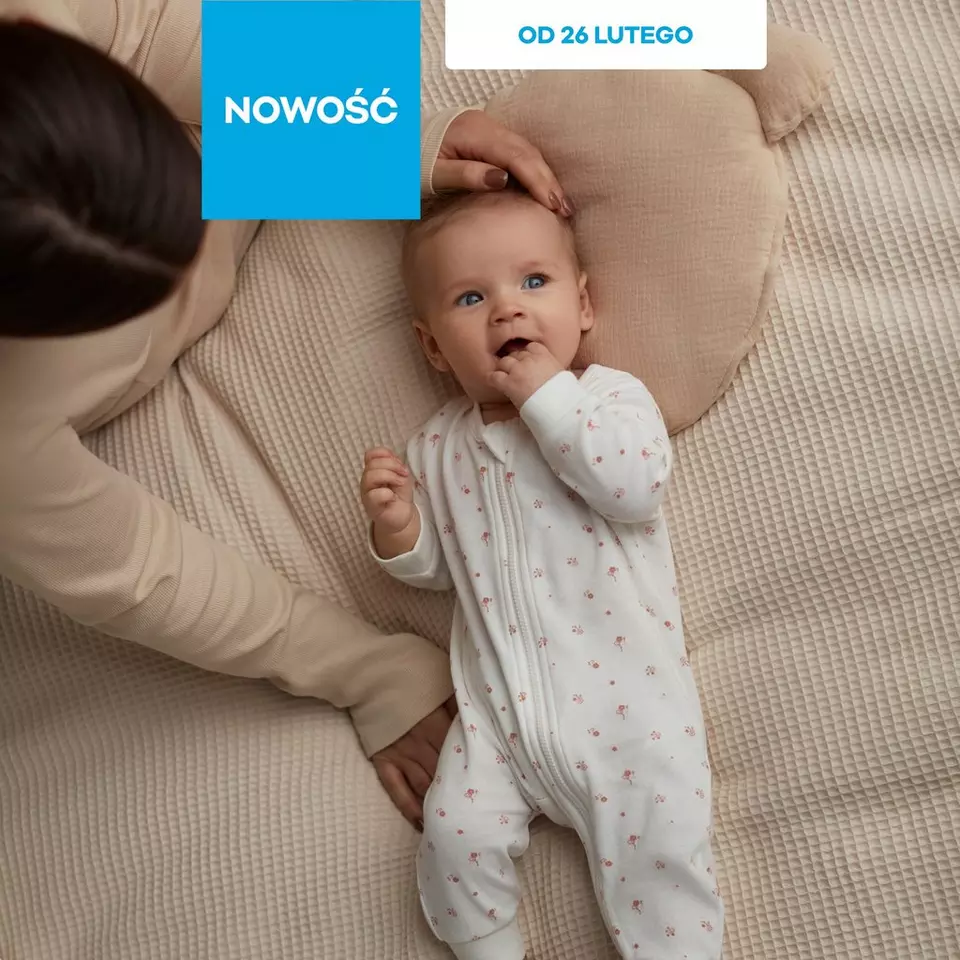 Niemowlę w białym śpioszkach z drobnymi różowymi wzorami, leżące na beżowej tkaninie. Napis "NOWOŚĆ" i "OD 26 LUTEGO".