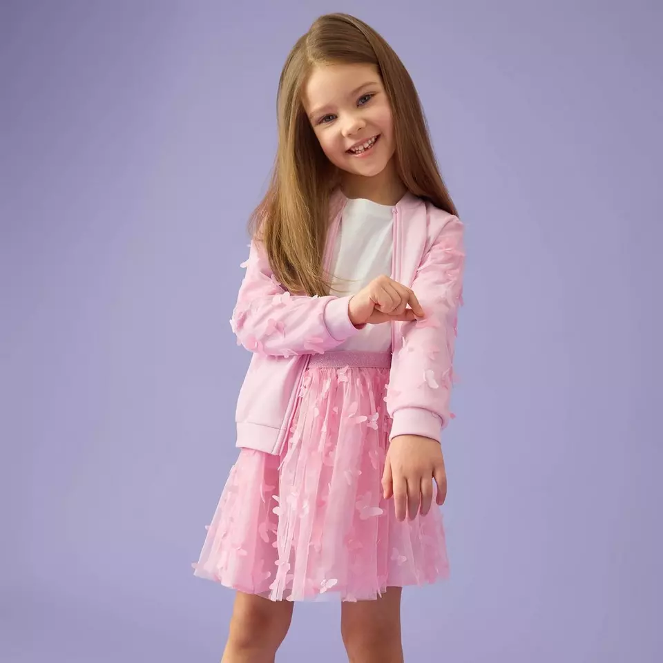 Young girl smiling wearing a pink satin bomber jacket and pink tulle skirt with butterfly details.