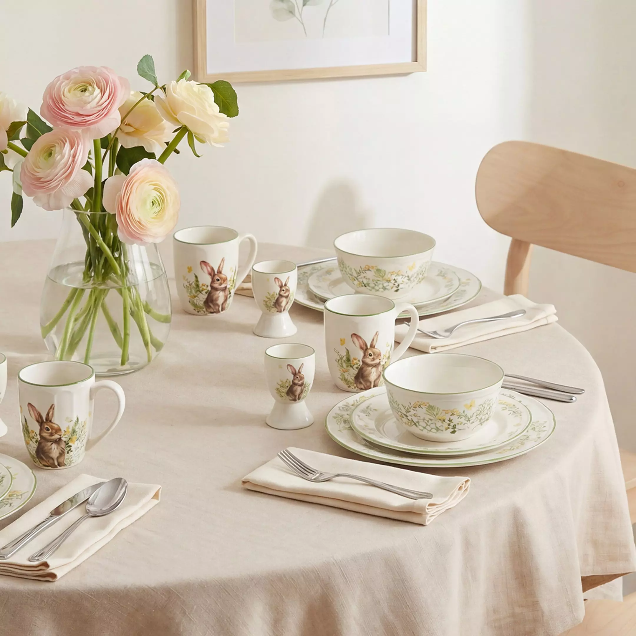 Easter dining set with rabbit-decorated mugs, floral bowls and plates on a cream tablecloth with pink ranunculus flowers in a vase.