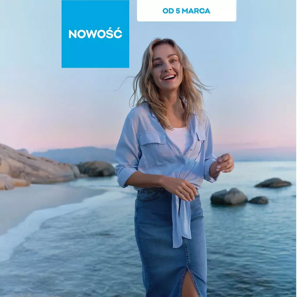 Uśmiechnięta kobieta w niebieskiej koszuli i dżinsowej spódnicy stoi nad morzem o zachodzie słońca.