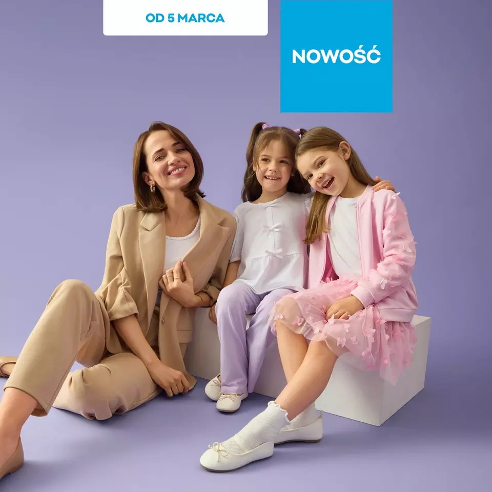 Kobieta w beżowym garniturze siedzi z dwiema dziewczynkami w pastelowych strojach, uśmiechając się.