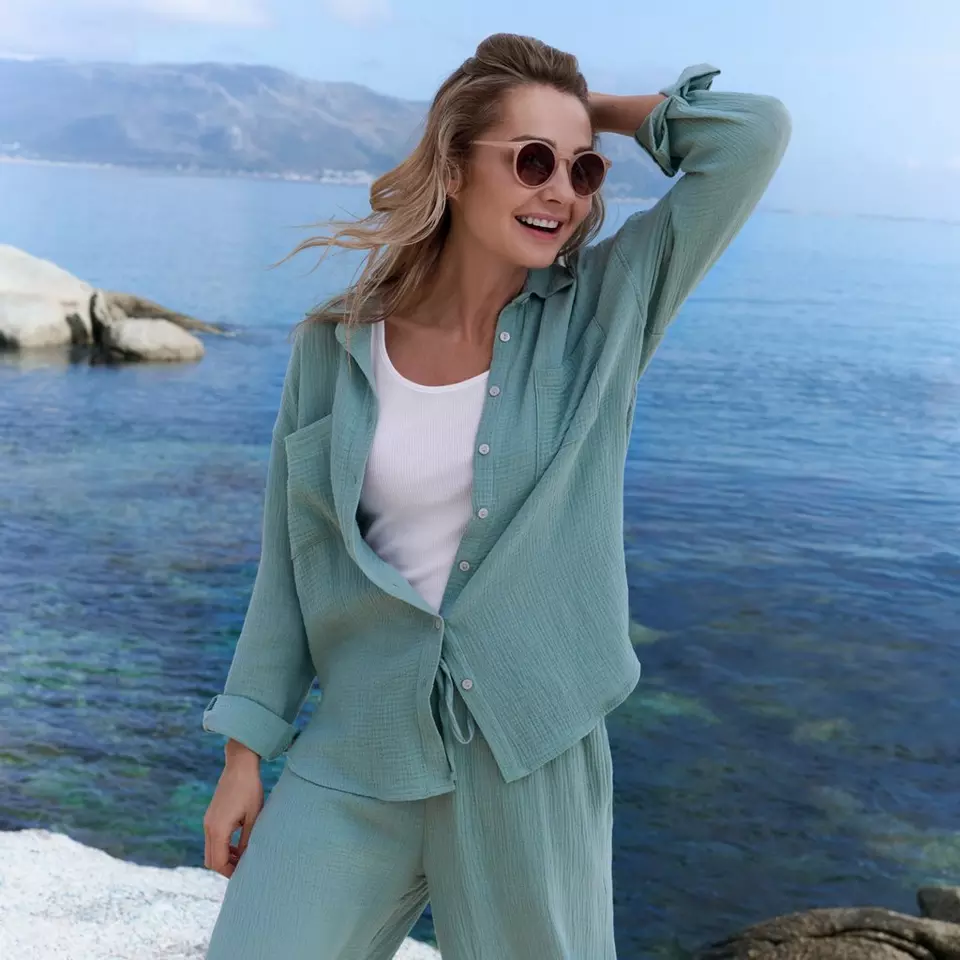 Woman wearing mint green textured shirt and trousers with round sunglasses by coastal water.