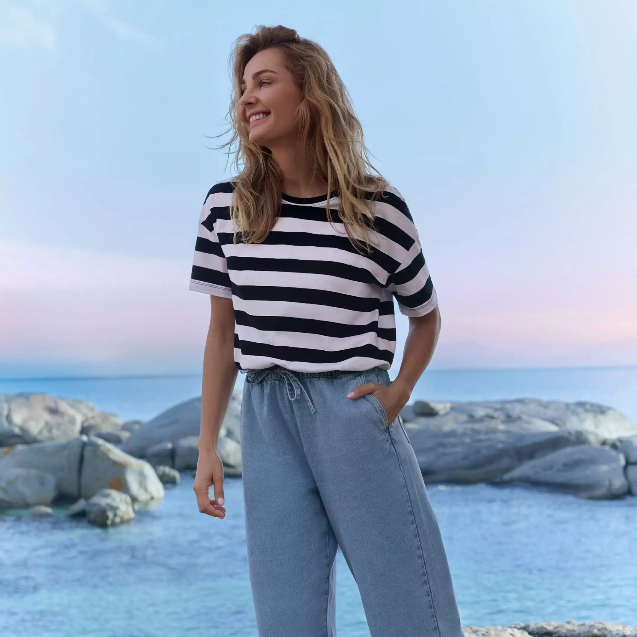 Woman smiling by the sea wearing black and white striped t-shirt with light blue denim trousers.
