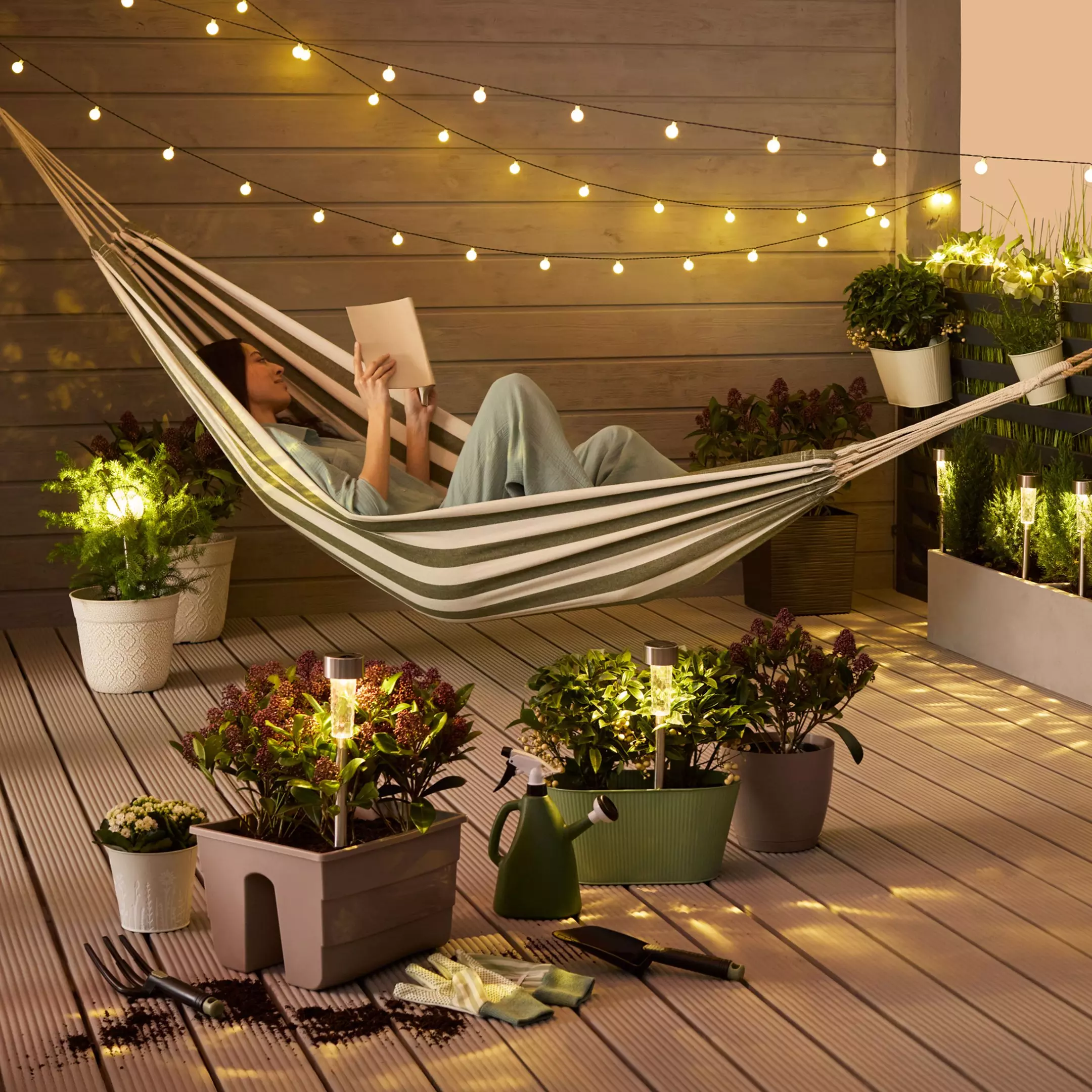 Person relaxing in striped hammock reading a book on wooden deck surrounded by plants and string lights.