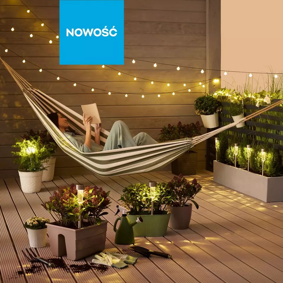 Osoba czytająca książkę w pasiastym hamaku na tarasie z roślinami i lampkami dekoracyjnymi.