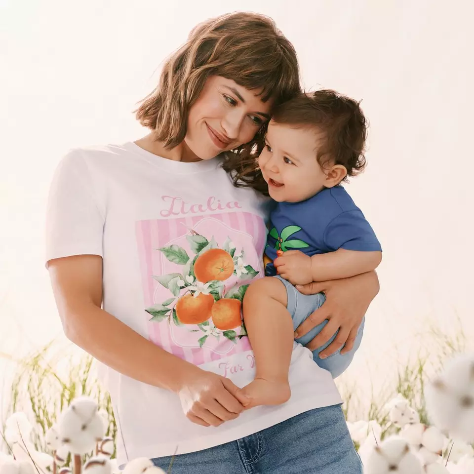 Mujer sonriente sosteniendo a un niño pequeño, viste camiseta blanca con estampado de naranjas Italia.