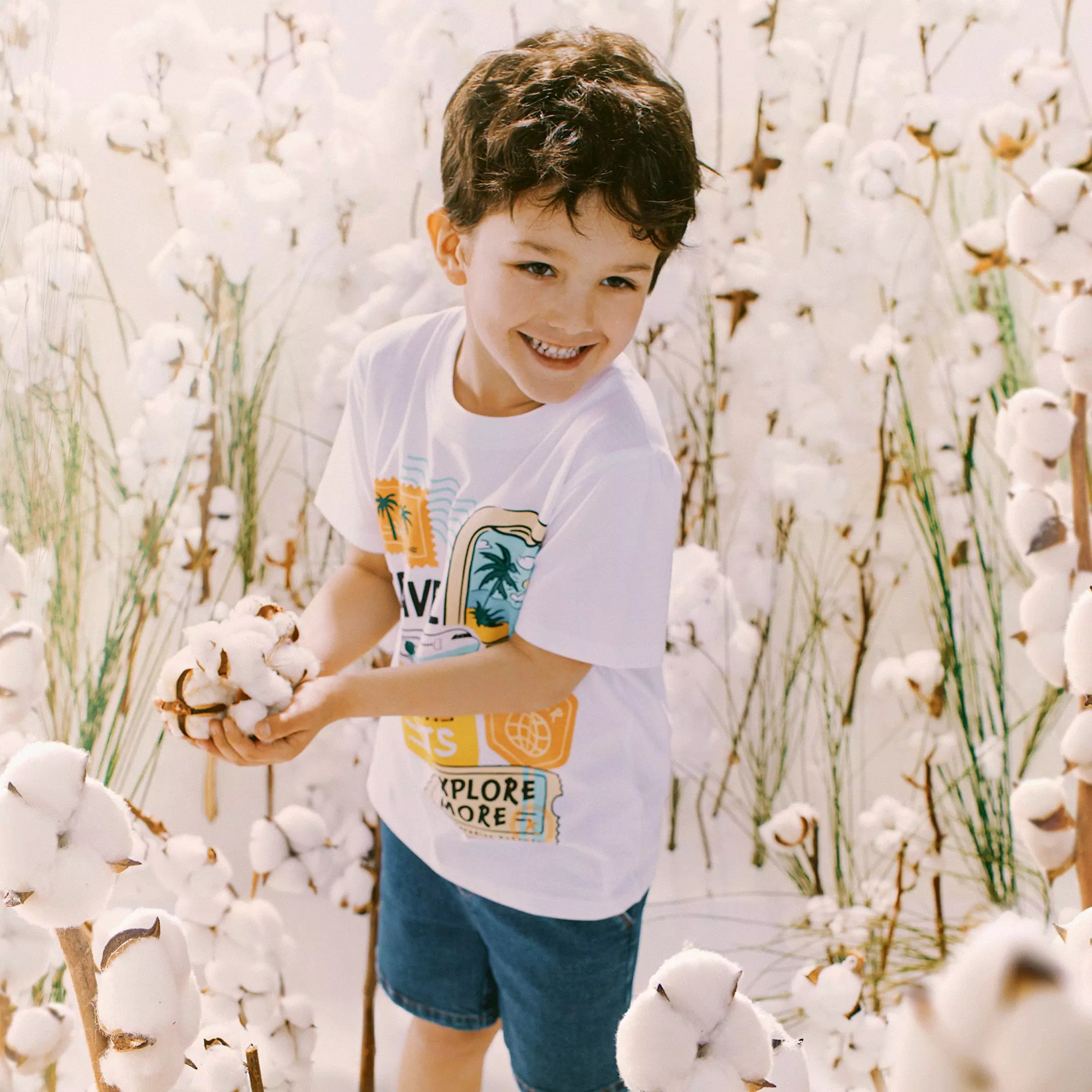 Niño sonriente sosteniendo flores de algodón en un campo de algodón, vistiendo camiseta blanca.