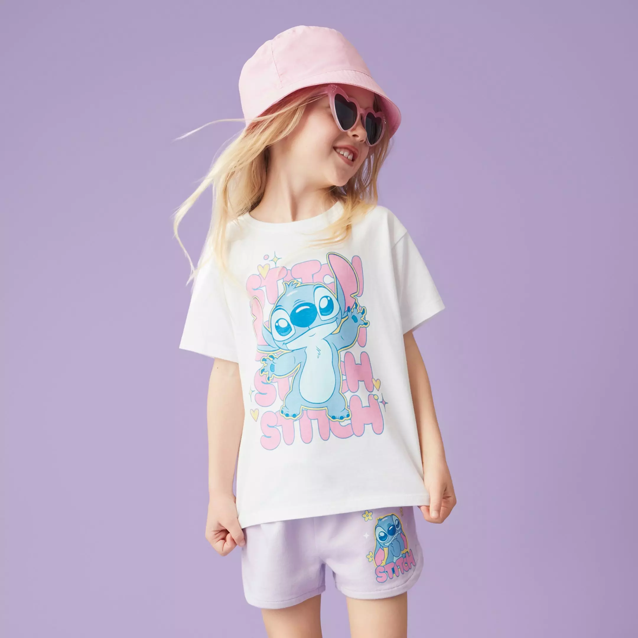 Fetiță purtând tricou alb cu Stitch, pălărie roz și ochelari de soare în formă de inimă.