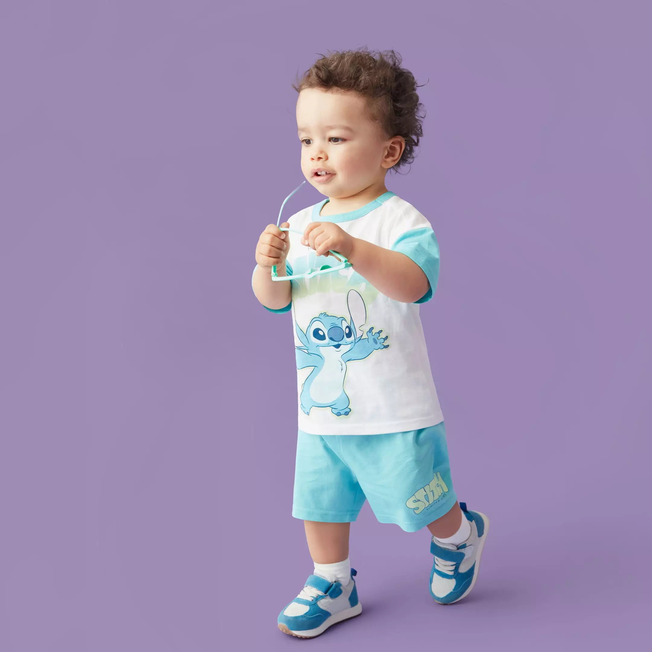 Copil mic purtând tricou alb cu personajul Stitch, pantaloni scurți turcoaz și adidași.