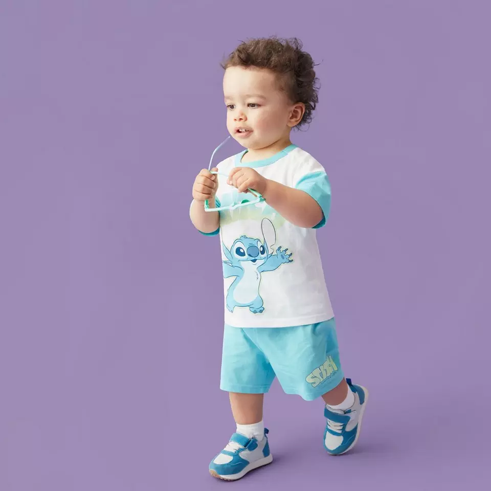 Copil mic purtând tricou alb cu personajul Stitch, pantaloni scurți turcoaz și adidași.