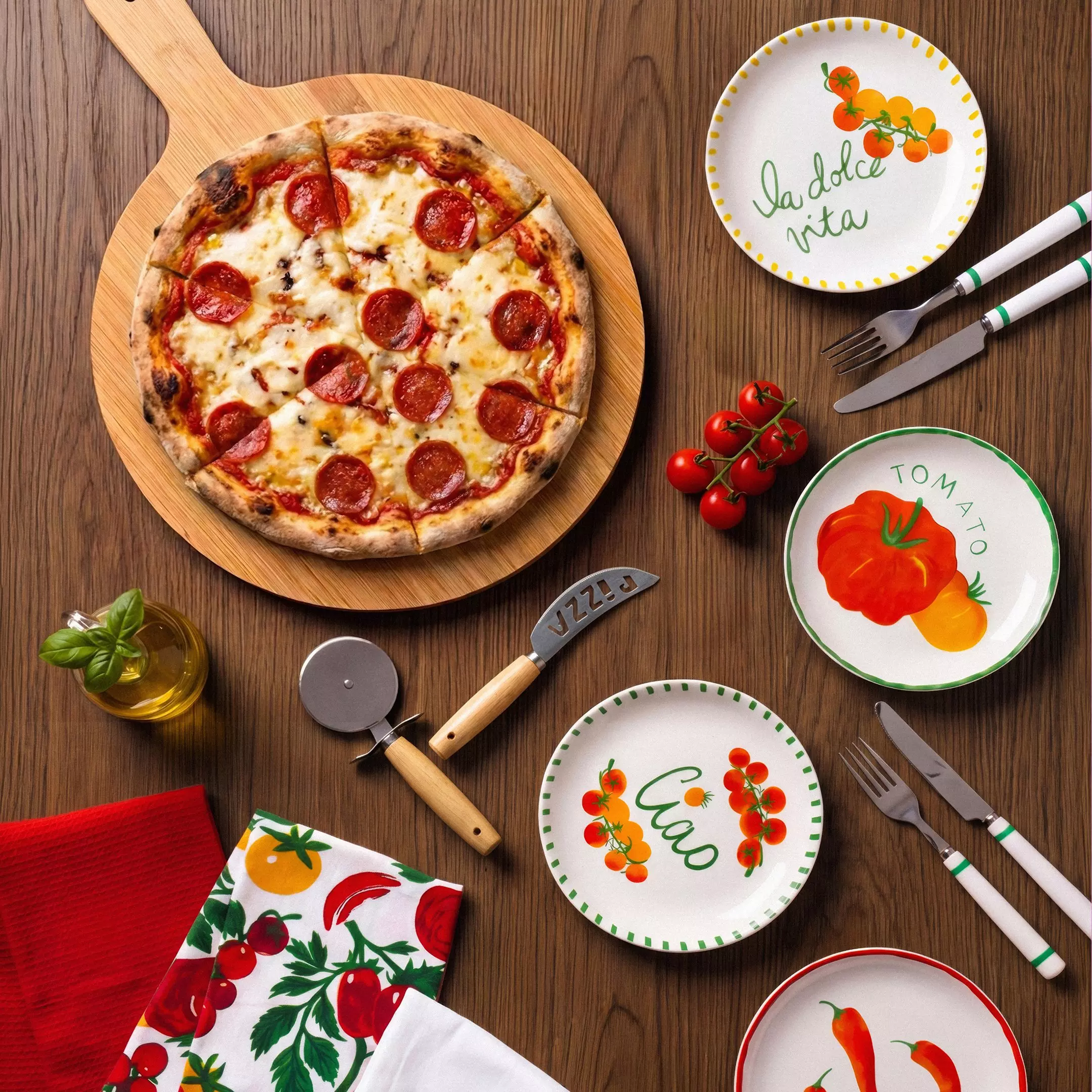 Pepco pepperoni pizza on wooden board with decorative Italian-themed plates, cutlery and fresh tomatoes.