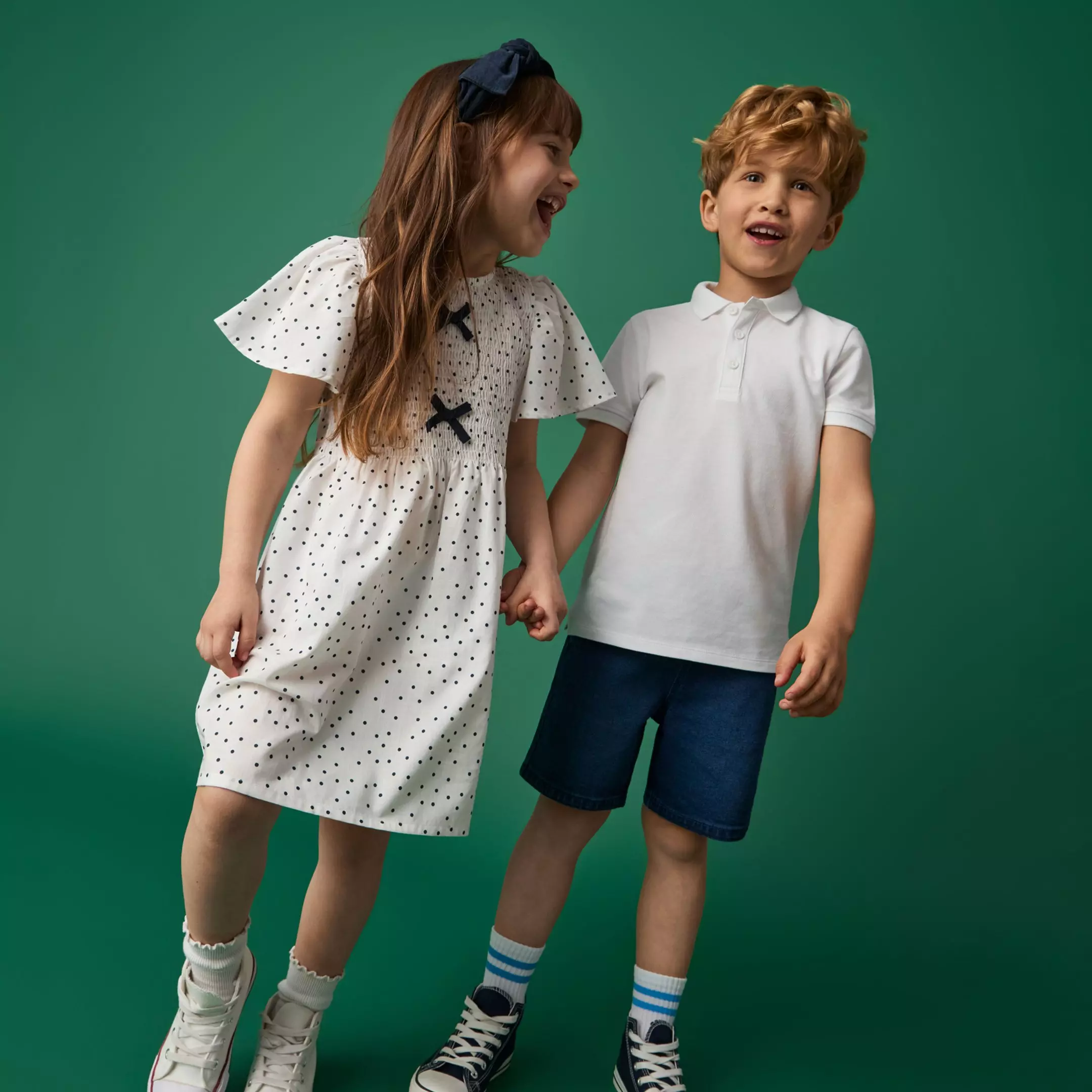 Niña con vestido blanco de lunares y niño con polo blanco sonriendo tomados de la mano, ropa infantil Pepco.