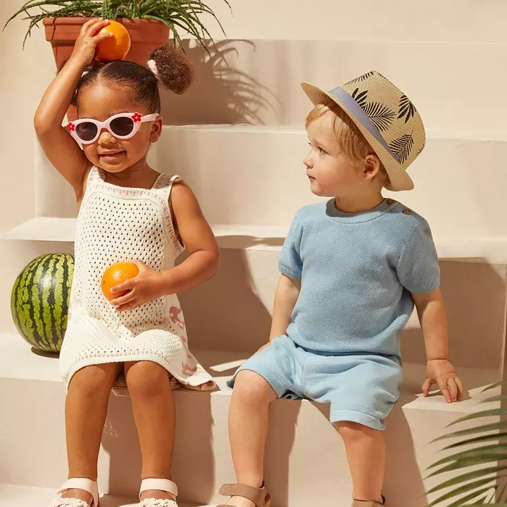Niños Pepco en ropa de verano jugando con frutas, una niña con gafas de sol y un niño con sombrero de paja.