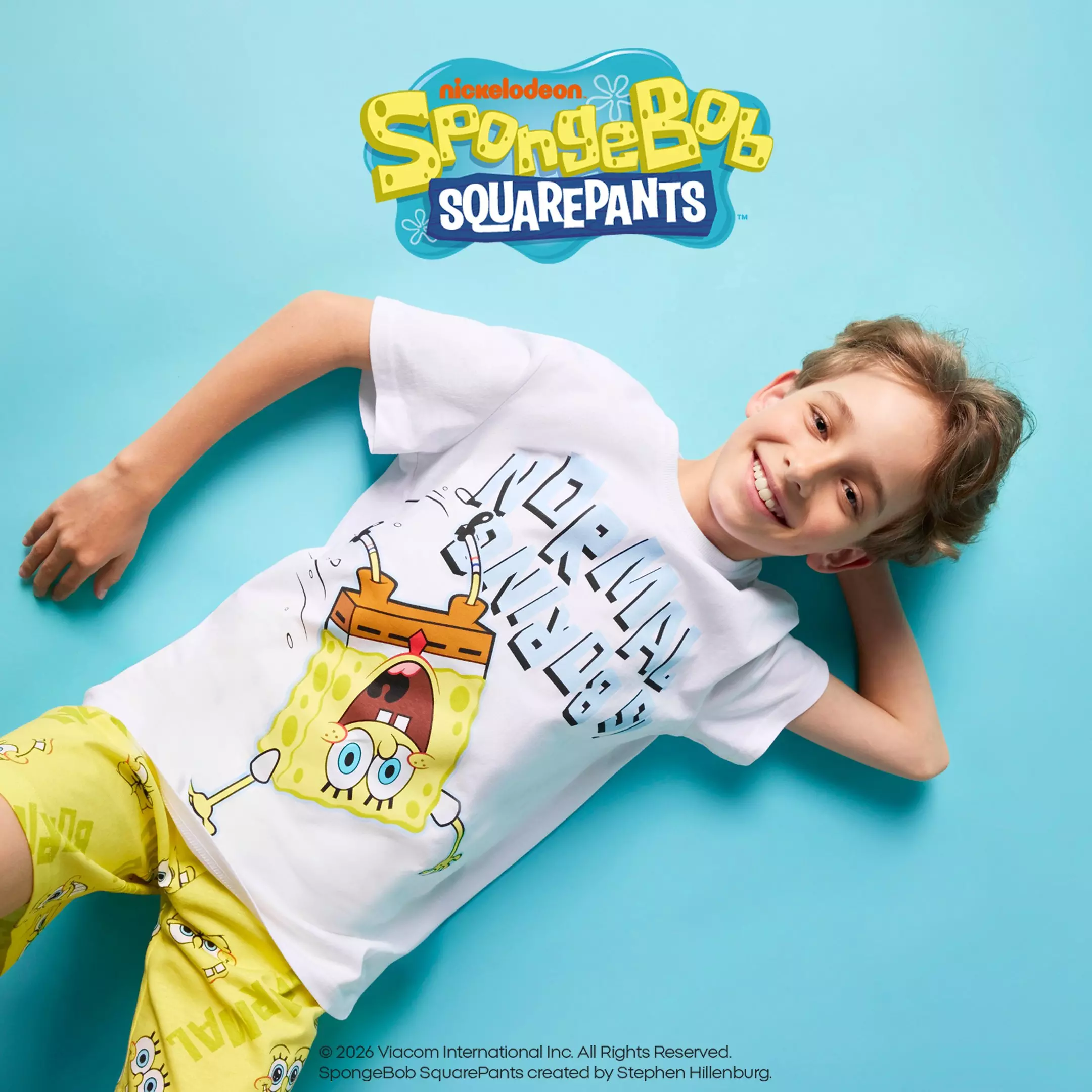 Chłopiec w białej koszulce Pepco z nadrukiem SpongeBob SquarePants leży uśmiechnięty na turkusowym tle.