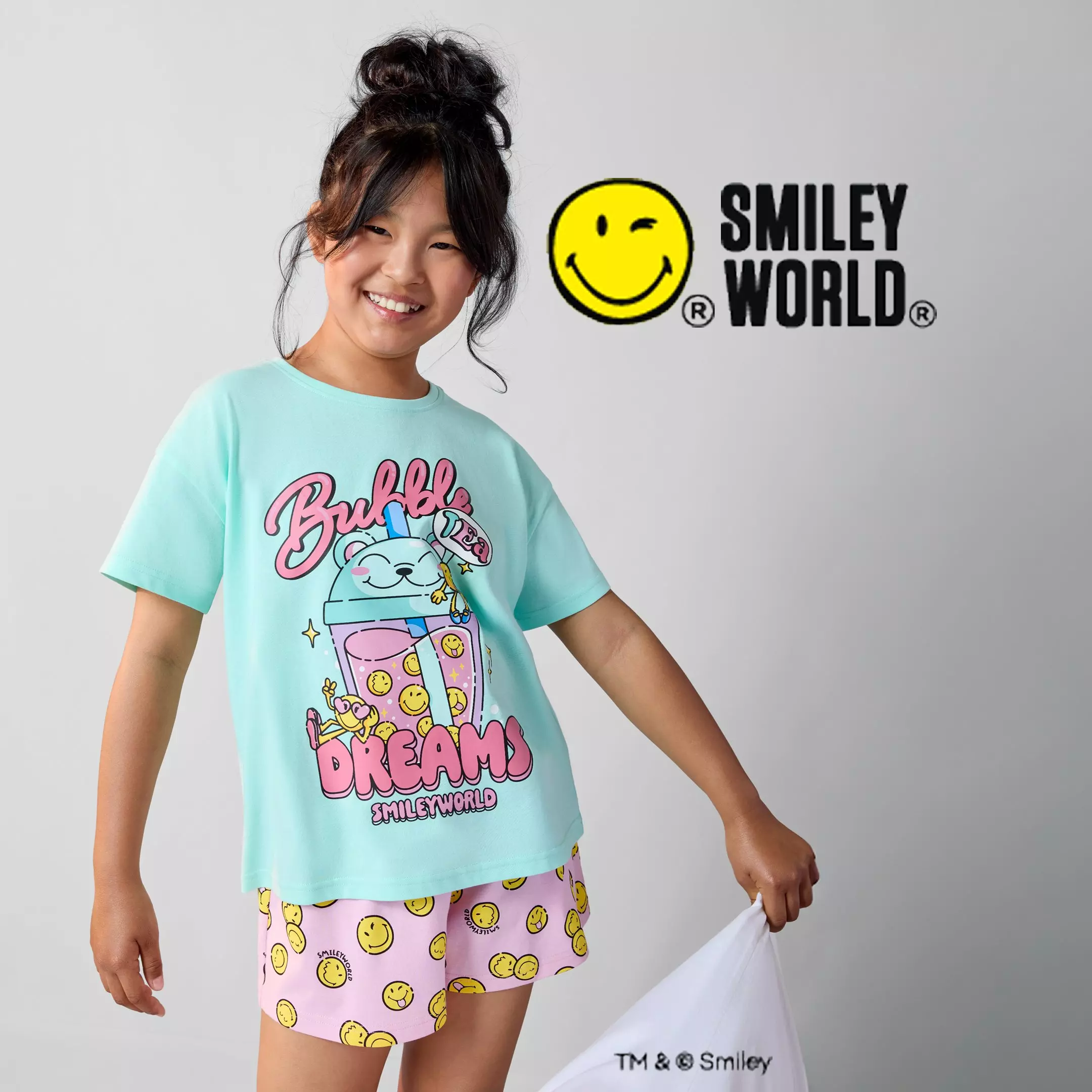 Dívka v tyrkysovém tričku a růžových šortkách Pepco s motivem SMILEY WORLD se usmívá.