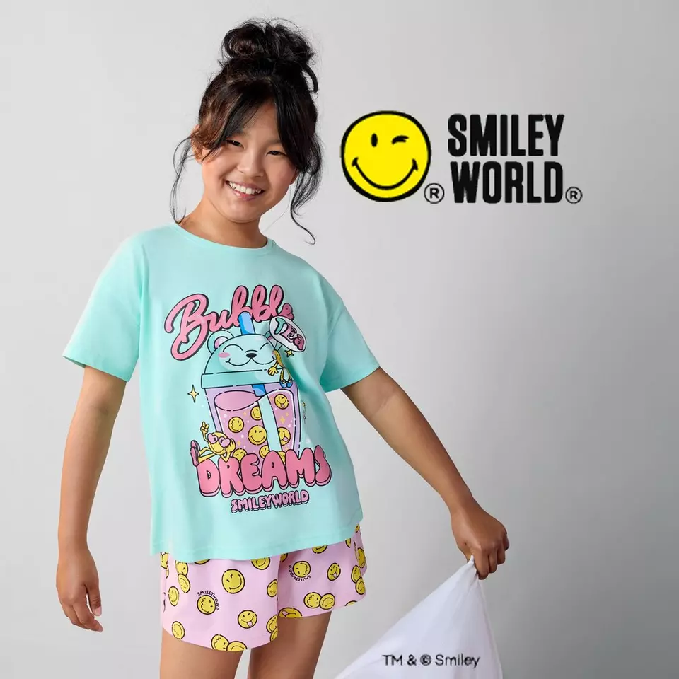 Dívka v tyrkysovém tričku a růžových šortkách Pepco s motivem SMILEY WORLD se usmívá.