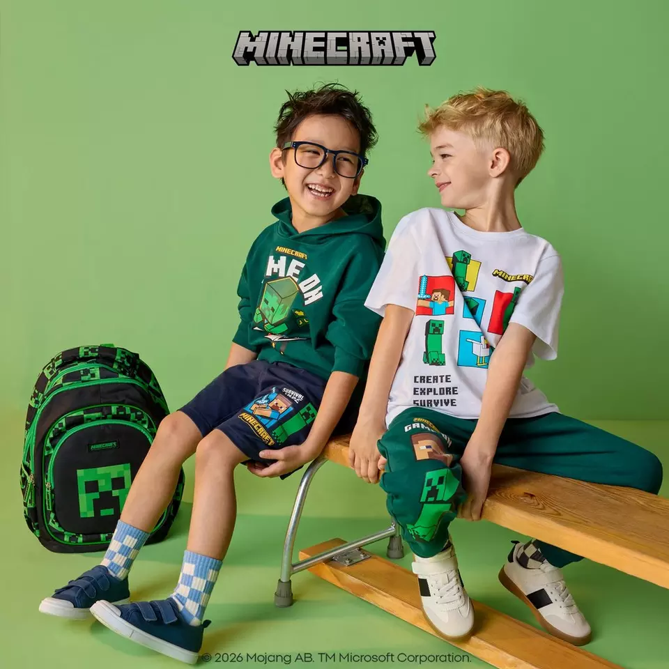 Doi băieți purtând îmbrăcăminte Pepco cu licență Minecraft, stând pe o banchetă cu un rucsac în apropiere.