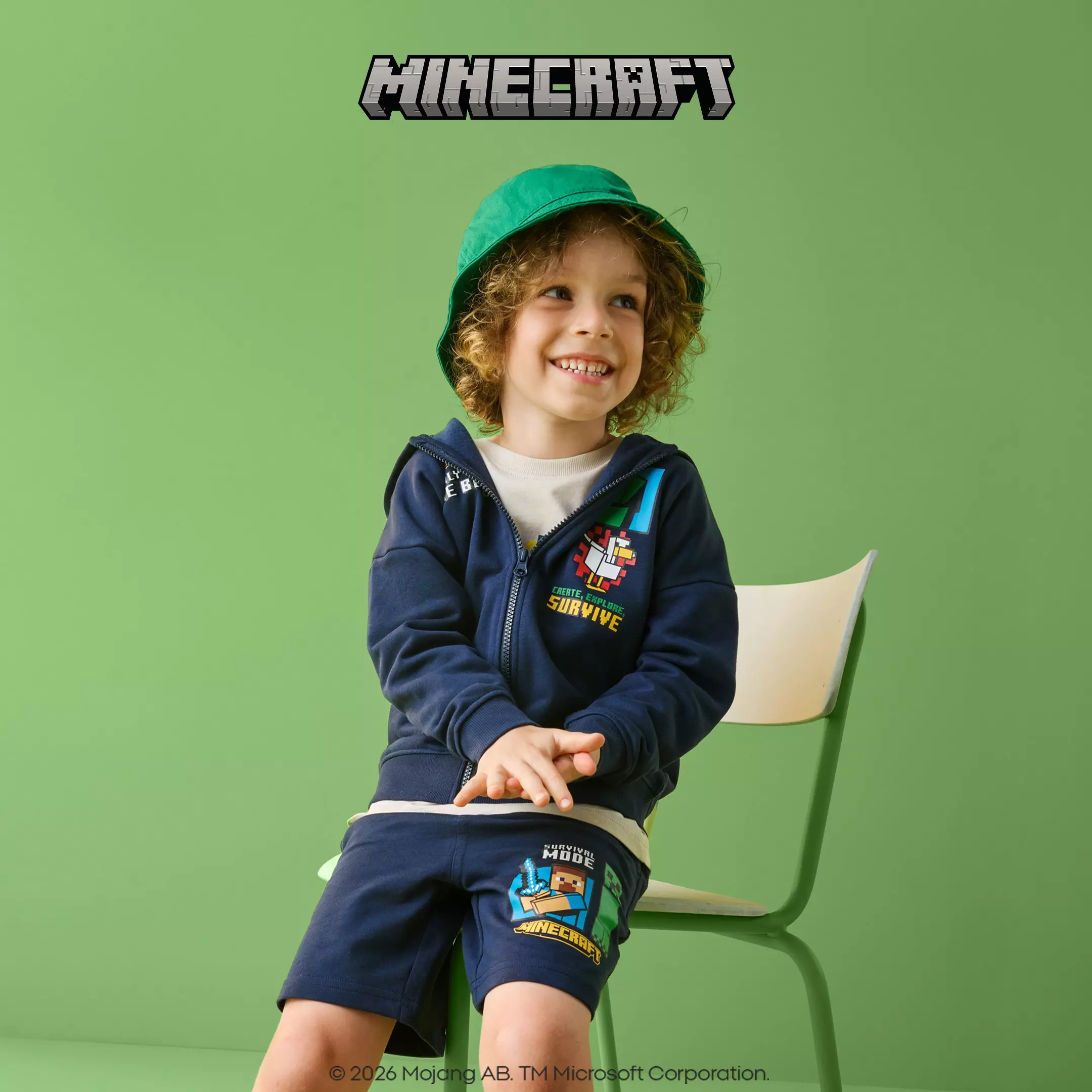 Pepco child wearing Minecraft branded navy hoodie and shorts with green bucket hat, sitting and smiling.