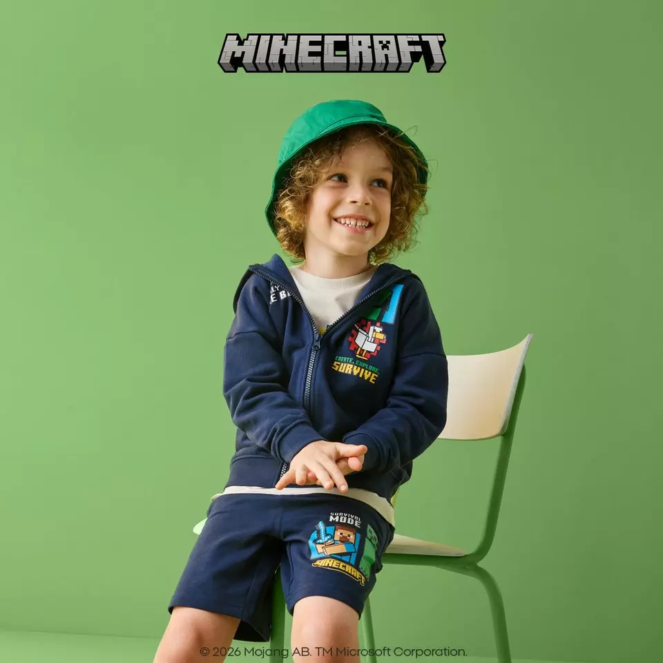 Pepco child wearing Minecraft branded navy hoodie and shorts with green bucket hat, sitting and smiling.