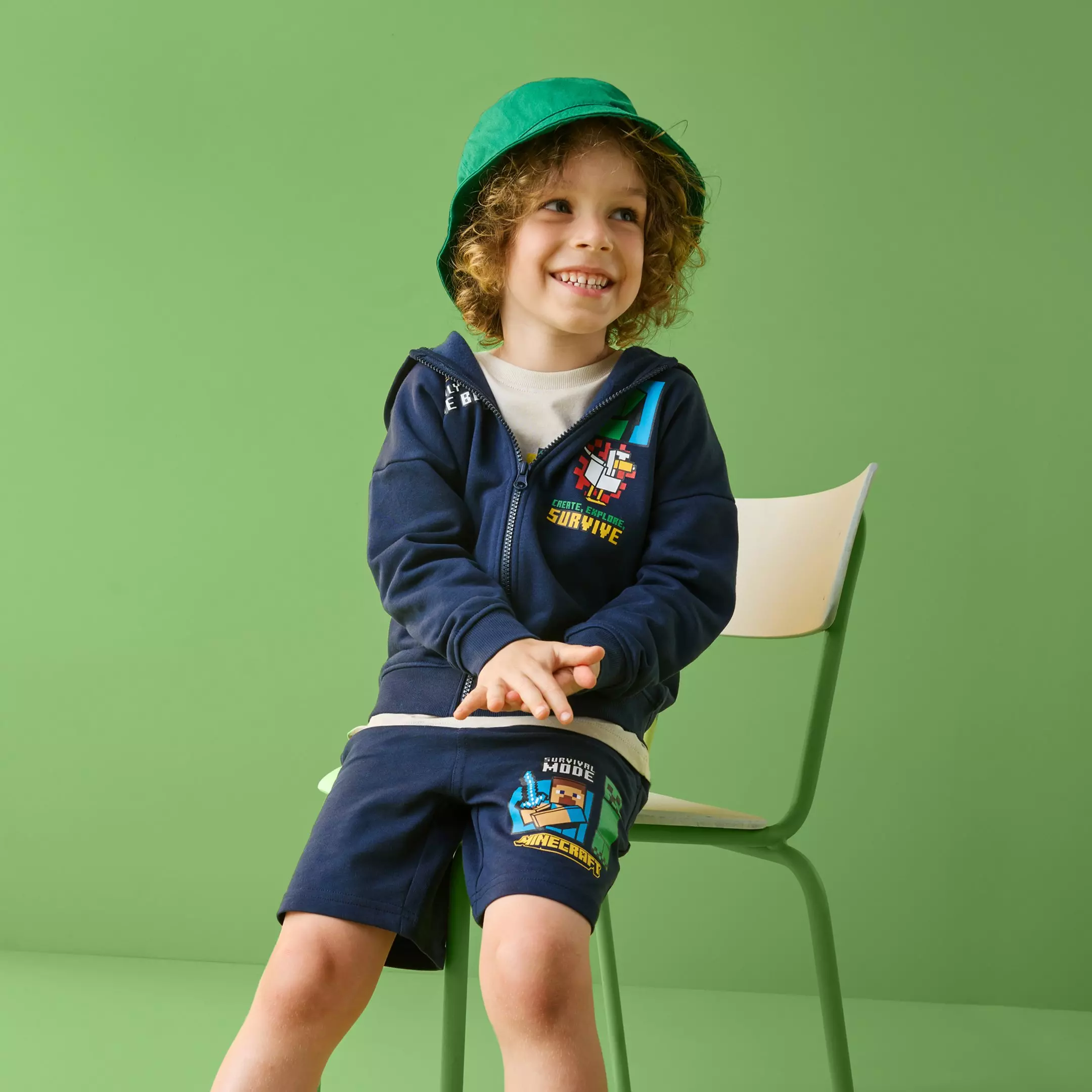 Băiat zâmbitor purtând set Pepco Minecraft cu hanorac bleumarin și pantaloni scurți, cu pălărie verde.