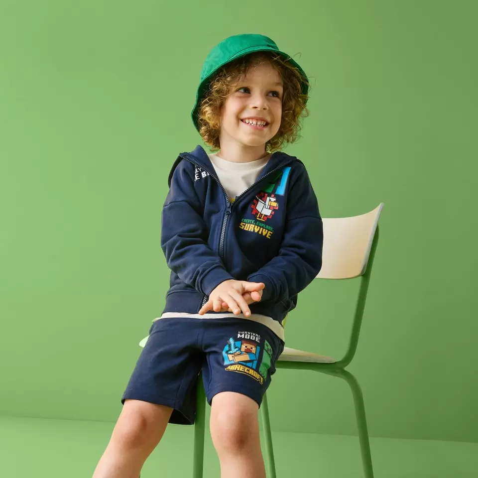 Băiat zâmbitor purtând set Pepco Minecraft cu hanorac bleumarin și pantaloni scurți, cu pălărie verde.