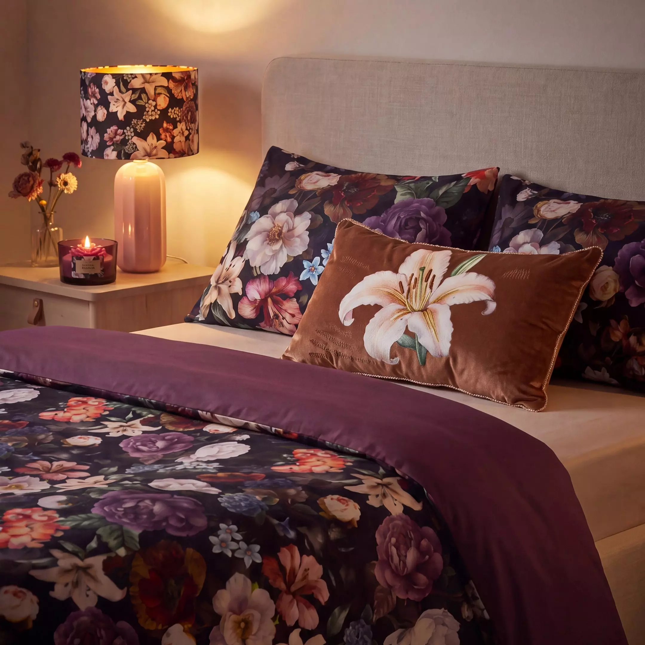 Lenjerie de pat Pepco cu model floral întunecat, perne decorative și lampă cu abajur asortată pe noptieră.