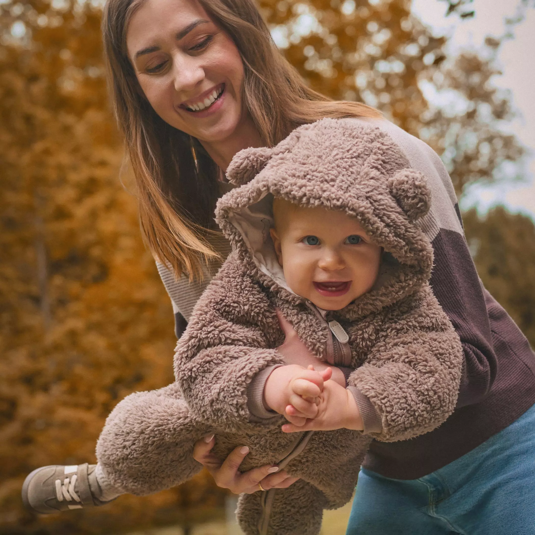 Madre sonriente sostiene a bebé que lleva mono de osito peludo marrón Pepco con capucha y orejas en otoño.