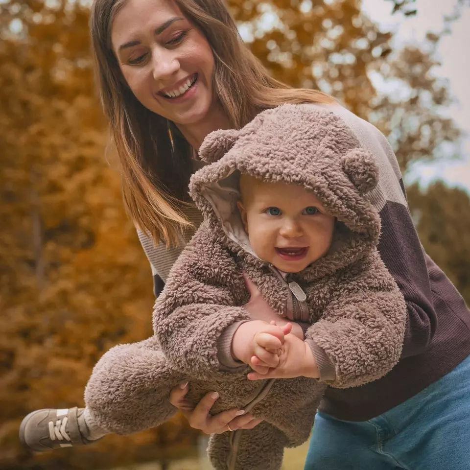 Madre sonriente sostiene a bebé que lleva mono de osito peludo marrón Pepco con capucha y orejas en otoño.