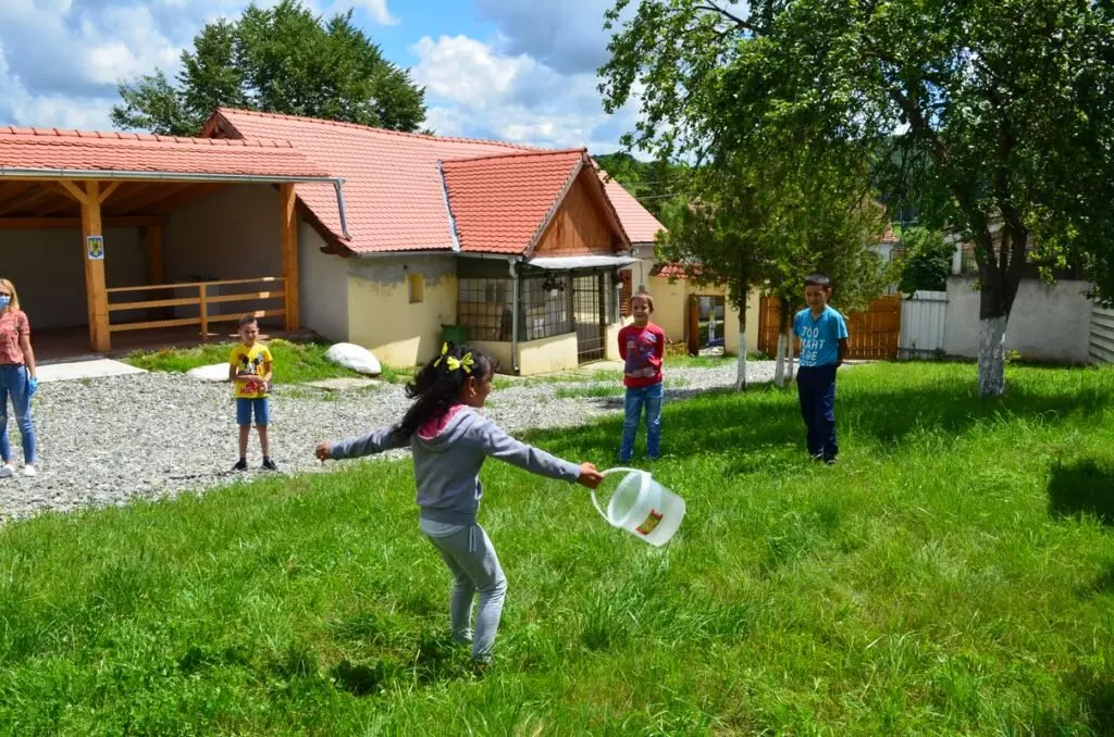 Copii jucându-se afară pe iarbă în fața unei clădiri cu acoperiș roșu într-o zi însorită.