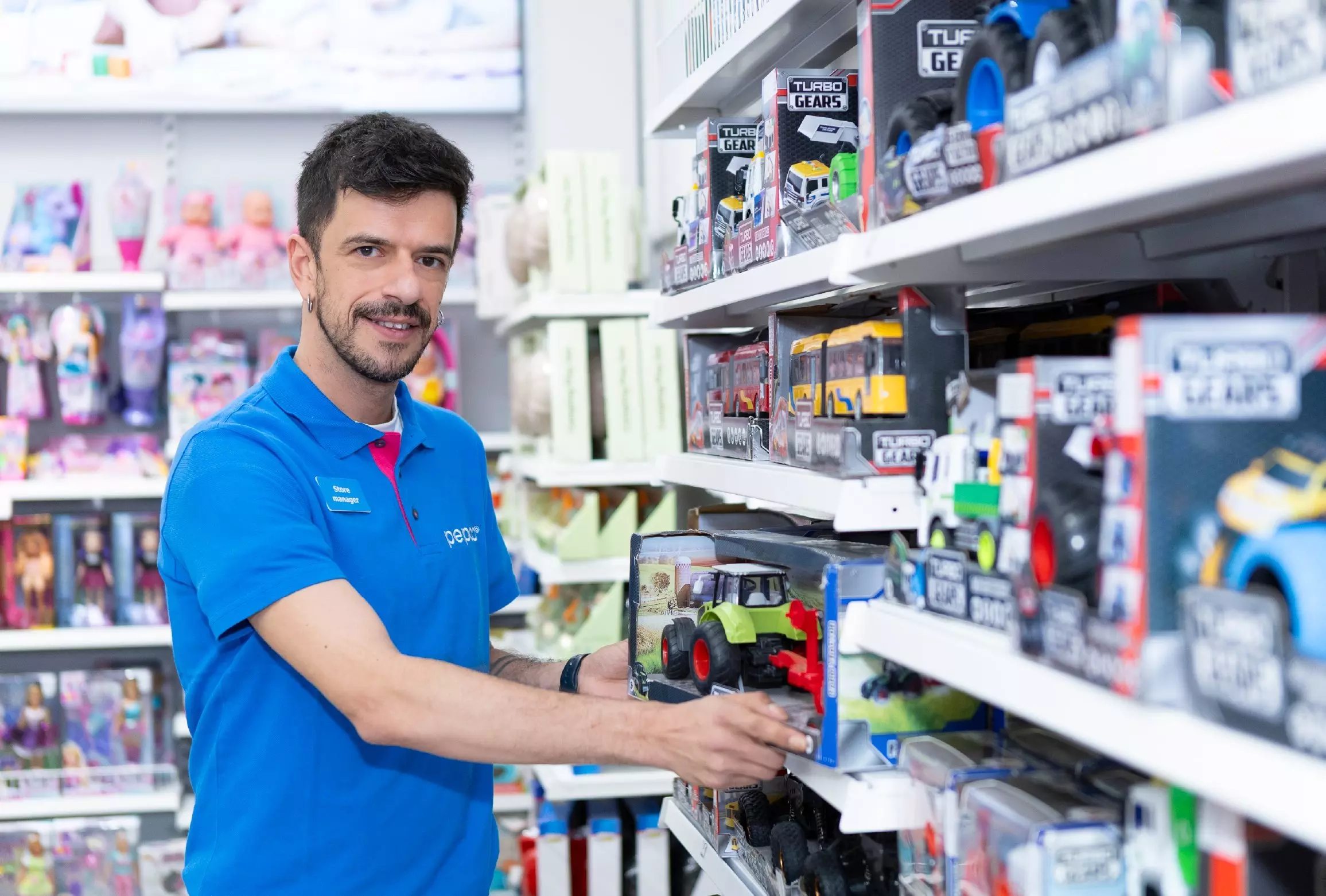 Angajat în tricou albastru aranjează jucării pe raft, ținând un tractor în cutie în magazin.