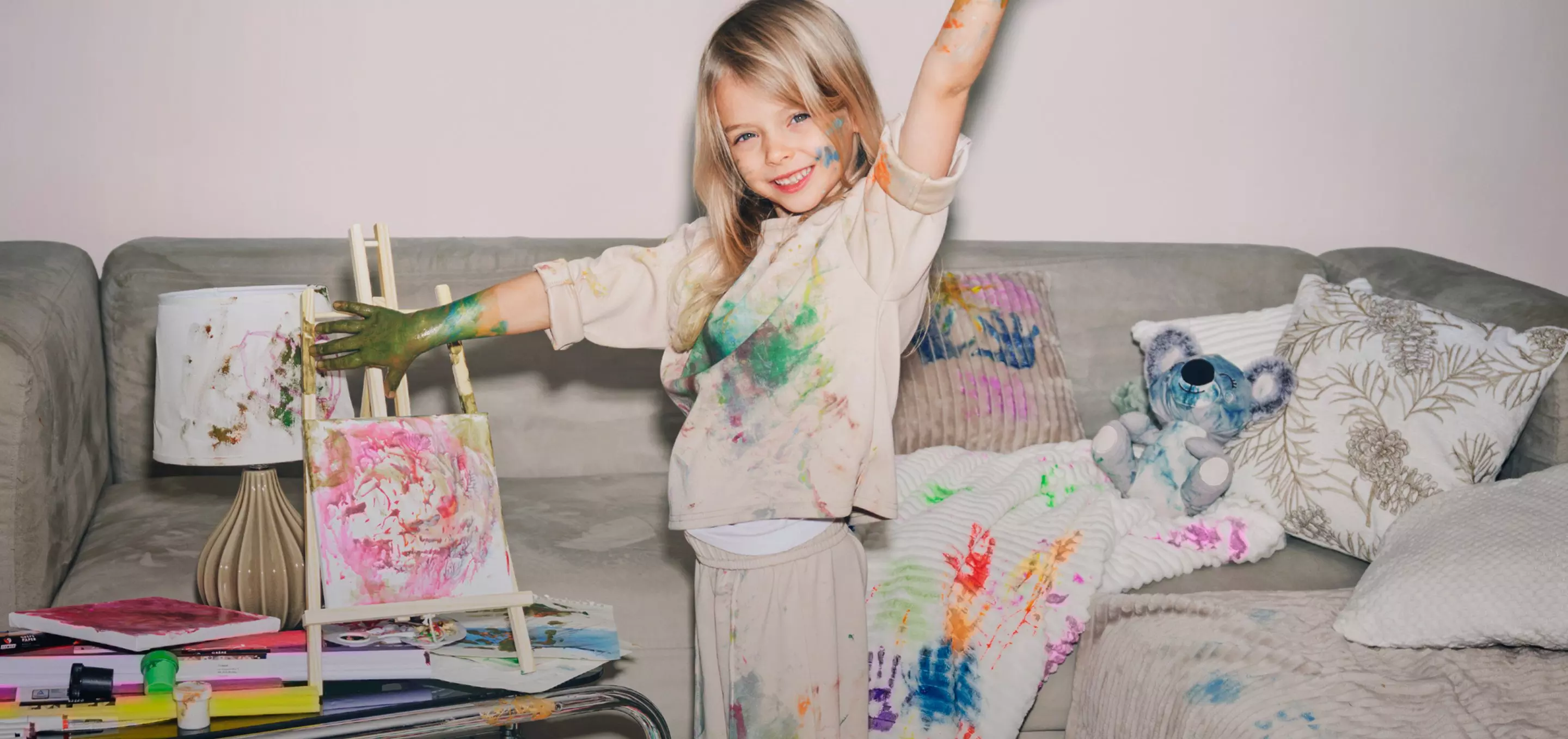 Dziecko w poplamionych farbą ubraniach maluje na sztaludze, stojąc na szarej kanapie z kolorowymi poduszkami.