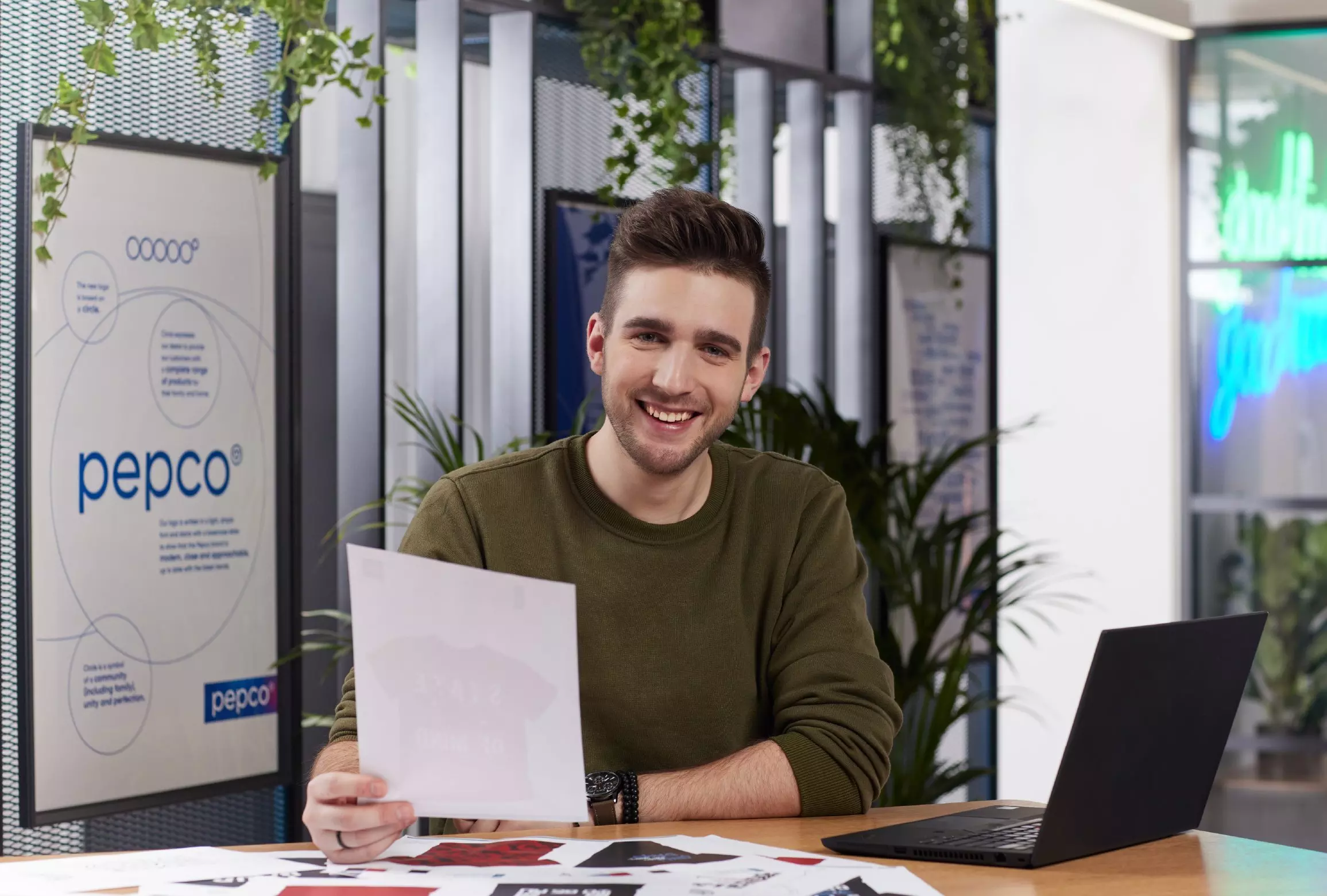 Bărbat zâmbitor în bluză verde ține un document alb, lucrând la laptop într-un birou modern.