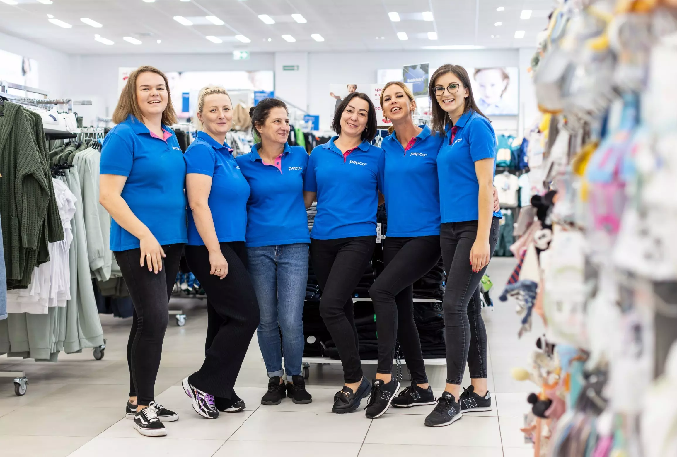 Șase angajate Pepco în uniforme albastre pozează zâmbind într-un magazin de retail.