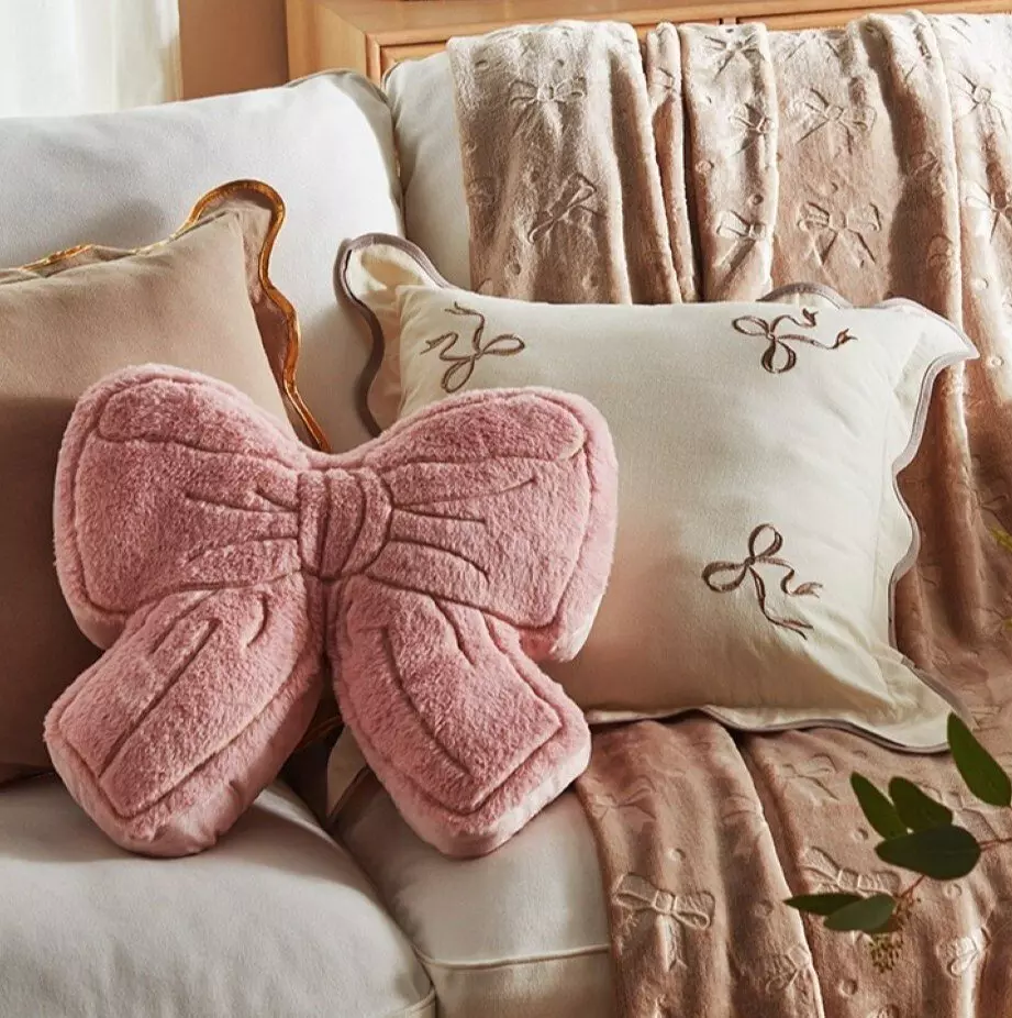 Różowa poduszka dekoracyjna Pepco w kształcie kokardki, obok kremowa poduszka z haftowanymi kokardkami i miękki koc.